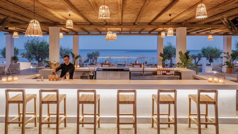 Beach Bar ong white counter with wooden stools, a bartender preparing cocktails, and a view of the ocean under a thatched roof with wicker pendant lights at Grecotel LUXME Dama Dama by Kenwood Travel
