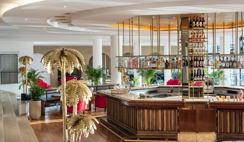 Lobby Bar gold palm tree sculptures, a well-stocked bar with brass accents, and bright, modern lounge seating at Grecotel LUXME Dama Dama by Kenwood Travel