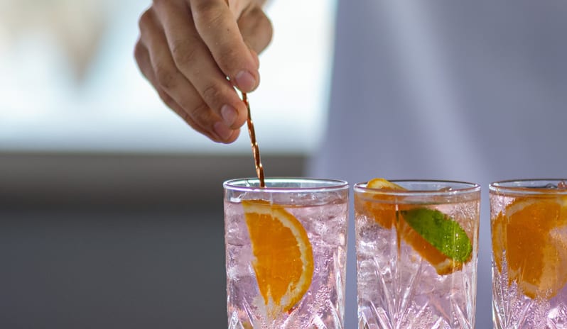 Beach Club Close-up of a bartender using a bar spoon to stir refreshing pink cocktails garnished with fresh orange and lime at Grecotel LUXME Dama Dama by Kenwood Travel