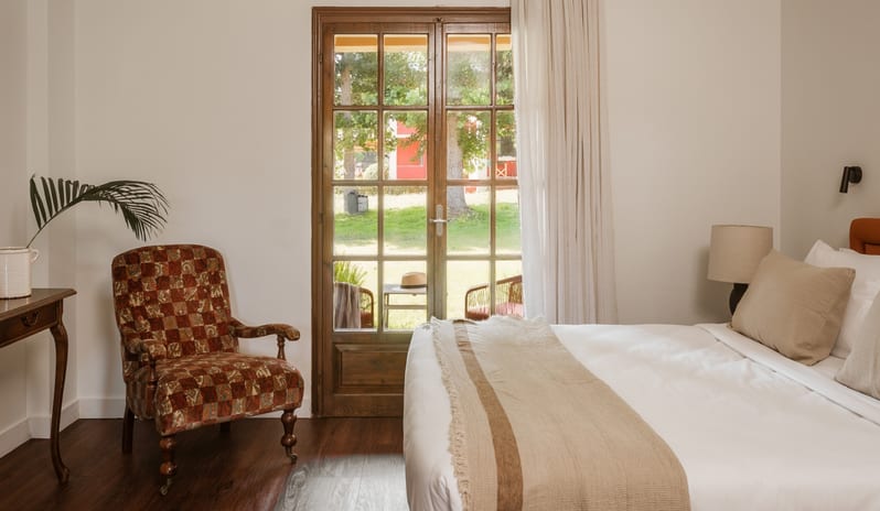 Botanica Room Private Balcony white bed with beige pillows, a vintage patterned armchair, and glass French doors opening to a green garden view at Grecotel LUXME Costa Botanica by Kenwood Travel