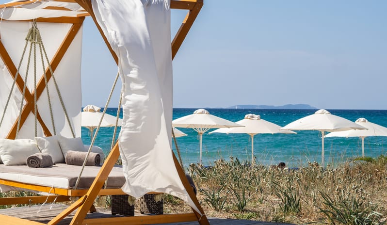 Beach Club A luxury wooden daybed with white cushions and flowing curtains at Grecotel LUXME Costa Botanica by Kenwood Travel