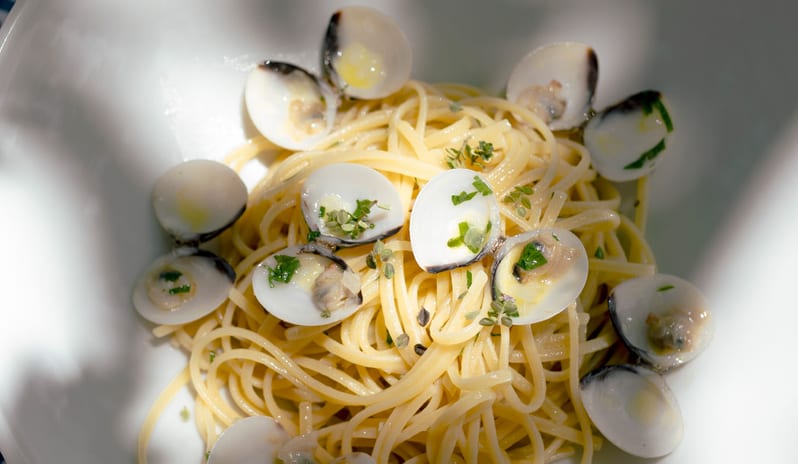 Nonna's A classic Italian linguine alle vongole (clam pasta) garnished with fresh herbs, presented in a deep white ceramic bowl on a patterned tablecloth under natural sunlight at Grecotel LUXME Costa Botanica by Kenwood Travel