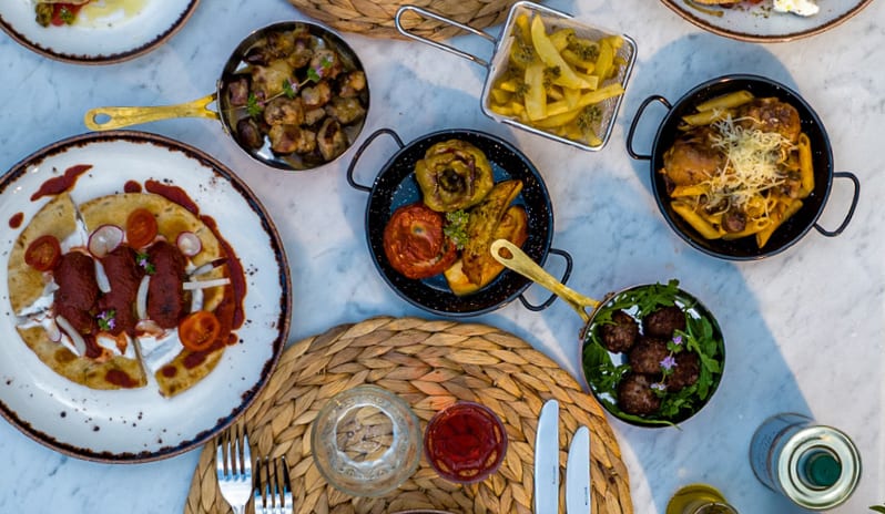 Tavernaki Flat lay of a traditional Greek meze feast on a white marble table with woven placemats, blue ceramic plates, fresh Greek salad, souvlaki, and various shared appetizers at Grecotel LUXME Costa Botanica by Kenwood Travel