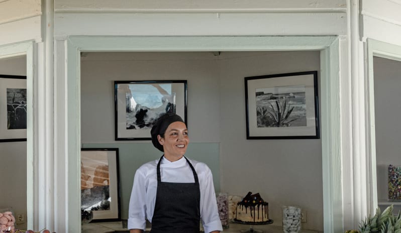 Beach Club A female pastry chef in a black apron and white uniform smiling at an outdoor resort dessert counter with fresh fruit and cakes at Grecotel LUXME Costa Botanica by Kenwood Travel