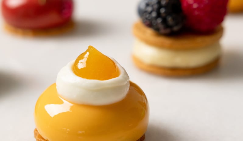 Patisserie & Chocolaterie Gourmet petit fours including a yellow mirror glaze mousse tart topped with cream and mango gelée alongside berry-topped cream biscuits at Grecotel LUXME Costa Botanica by Kenwood Travel