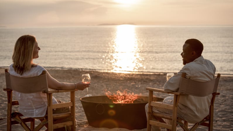 Beach Bar Man and woman sitting in director chairs drinking wine next to a fire pit on a beach in Corfu during sunset at Grecotel LUXME Costa Botanica by Kenwood Travel