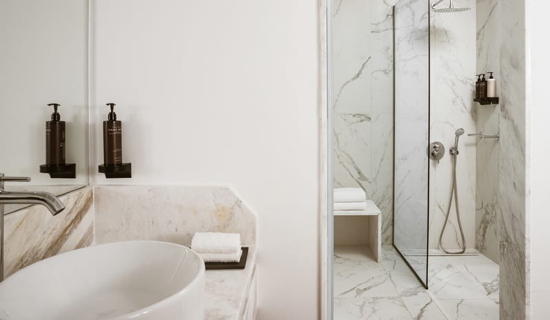 Family Apartment Private Balcony bathroom design with white marble floors and walls, a walk-in rainfall shower, folded white towels, and contemporary fixtures at Grecotel LUXME Costa Botanica by Kenwood Travel