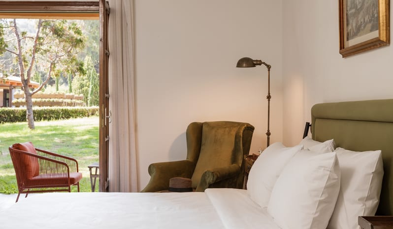 Botanica Room Private Balcony white bed with beige pillows, a vintage patterned armchair, and glass French doors opening to a green garden view at Grecotel LUXME Costa Botanica by Kenwood Travel