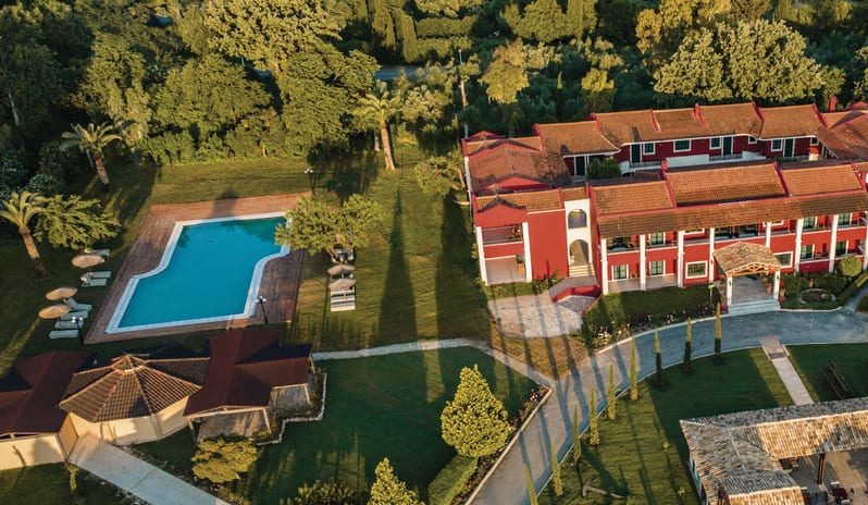 Grecotel LUXME Costa Botanica Family Suite at Yuccas Residence Pr Balcony sharing Pool High-angle view of a Mediterranean resort in Greece with a red estate building, outdoor pool, sun loungers, and surrounding forest landscape at sunset-Kenwood Travel