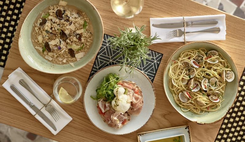 Nonna's Overhead view of an Italian restaurant table setting featuring a truffle mushroom risotto, linguine alle vongole (clam pasta), and a prosciutto burrata bruschetta served with white wine at Grecotel LUXME Costa Botanica by Kenwood Travel