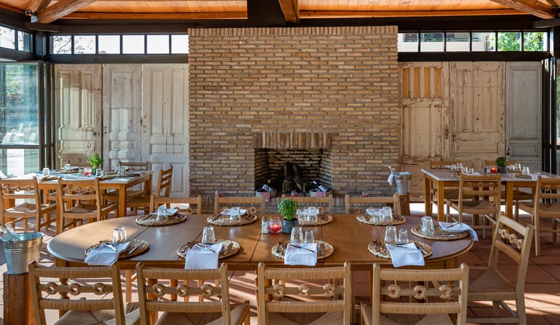 Tavernaki Rustic Mediterranean restaurant interior featuring a large exposed brick fireplace centerpiece, distressed wooden doors as wall panels, and woven rush-seat chairs at Grecotel LUXME Costa Botanica by Kenwood Travel