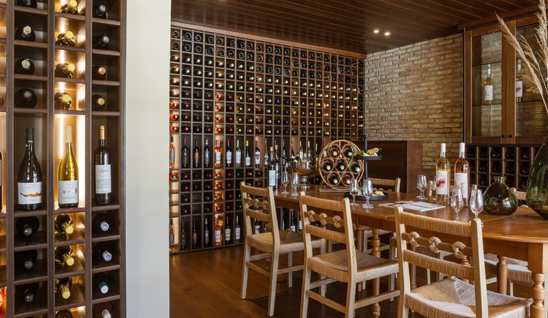 Botegga Cava di Vino custom wooden wine storage, rustic brick accents, and a tasting table with woven chairs at Grecotel LUXME Costa Botanica by Kenwood Travel
