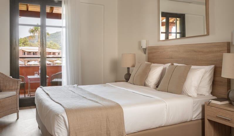 Botanica Bungalow Loft Private Balcony Modern Mediterranean style bedroom with neutral tones, light wood headboard, beige throw pillows, and glass balcony doors letting in natural light at Grecotel LUXME Costa Botanica by Kenwood Travel
