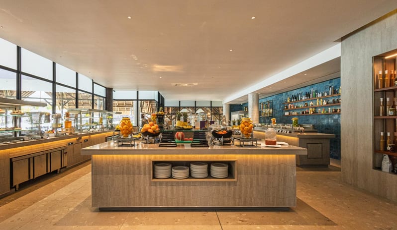 Le Grand Port Modern luxury hotel buffet area with fruit displays, warm lighting, and large windows at Shandrani for 2 by Kenwood Travel