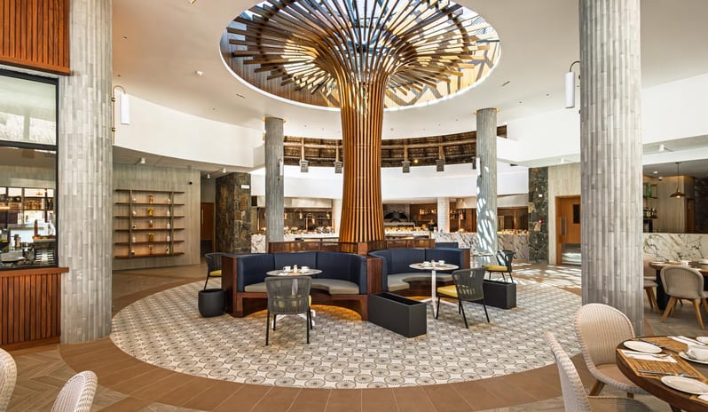 Le Grand Port Luxury resort restaurant dining area with architectural wooden tree feature, skylight, patterned tile flooring, and round booths at Shandrani for 2 by Kenwood Travel