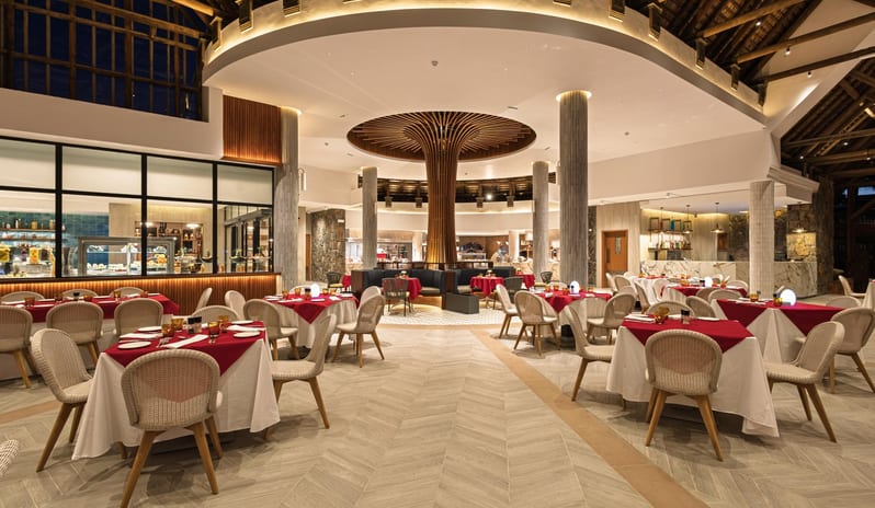 Le Grand Port Luxury resort restaurant interior at night featuring tables with red linens, an open kitchen, and a decorative wooden center column at Shandrani for 2 by Kenwood Travel