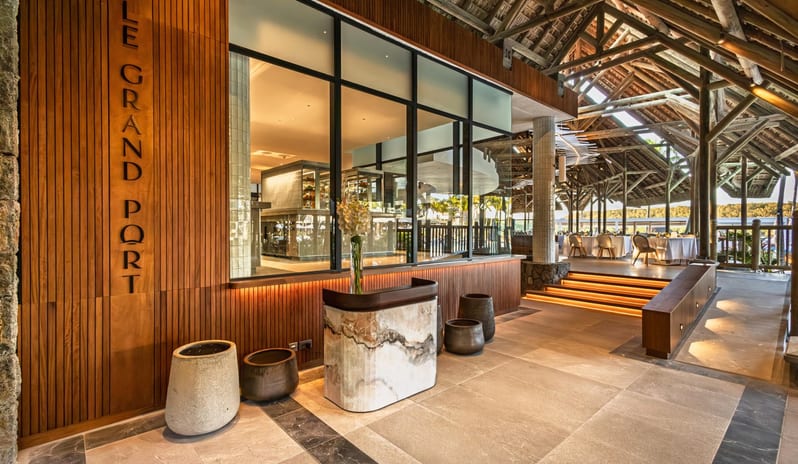 Le Grand Port vertical wood paneling, a marble reception desk, and a view into the open kitchen and thatched-roof outdoor dining area at Shandrani for 2 by Kenwood Travel