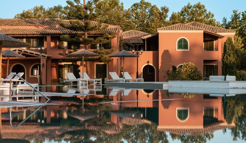 Botanica Bungalow Loft Private Balcony Terracotta-toned resort buildings with tiled roofs and arched doorways, perfectly reflected in a calm swimming pool lined with lounge chairs and straw umbrellas at Grecotel LUXME Costa Botanica by Kenwood Travel