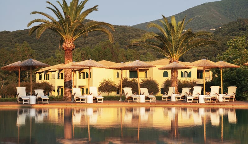 Family Loft Home Private Balcony Sunloungers and straw umbrellas reflecting in a luxury resort swimming pool with palm trees and a yellow villa in the background at Grecotel LUXME Costa Botanica by Kenwood Travel