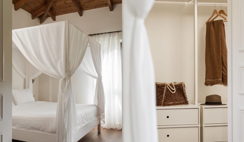 Family Loft Home Private Balcony Bright bohemian resort bedroom with a white canopy bed, exposed wood beam ceiling, and minimalist open closet at Grecotel LUXME Costa Botanica by Kenwood Travel