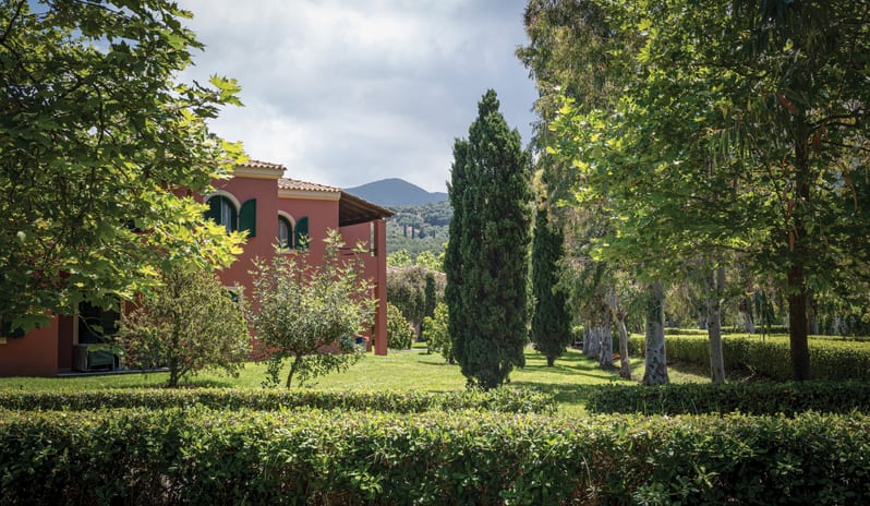 Play Room Suite Traditional Mediterranean-style estate nestled in lush green gardens with tall cypress trees and a mountain backdrop under a cloudy sky at Grecotel LUXME Costa Botanica by Kenwood Travel