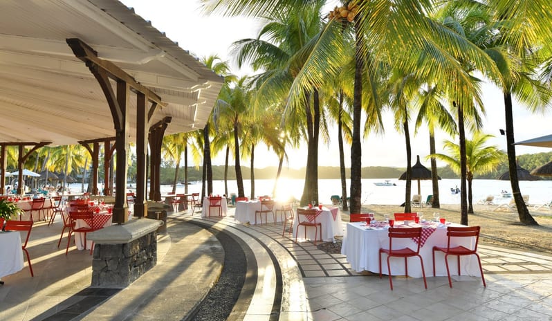 Ponte Vecchio Seaside restaurant tables on a paved patio overlooking a tropical beach with palm trees at Shandrani for 2 by Kenwood Travel