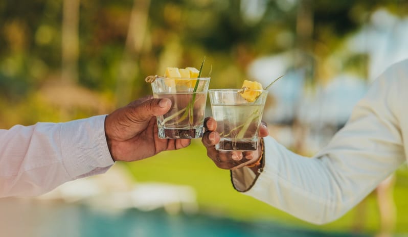 Purple for 2 Bar Two men clinking clear tropical cocktails garnished with pineapple chunks poolside at a luxury resort at Shandrani for 2 by Kenwood Travel