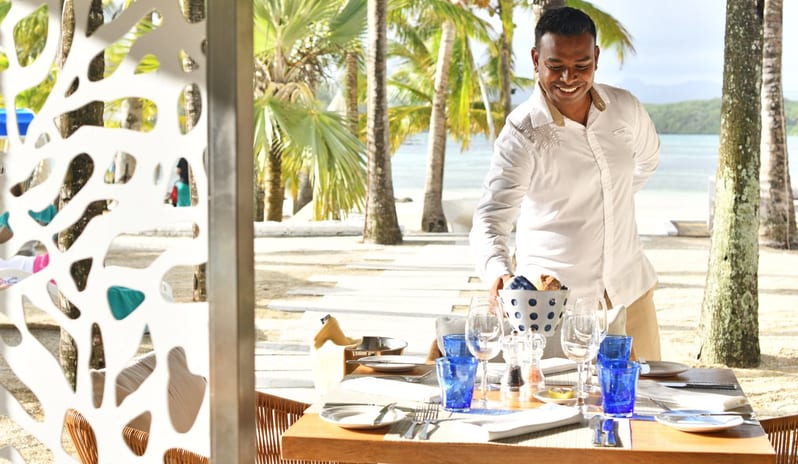 Le Sirius Wooden outdoor restaurant table with blue water glasses and a modern coral-patterned privacy screen in a tropical setting at Shandrani for 2 by Kenwood Travel