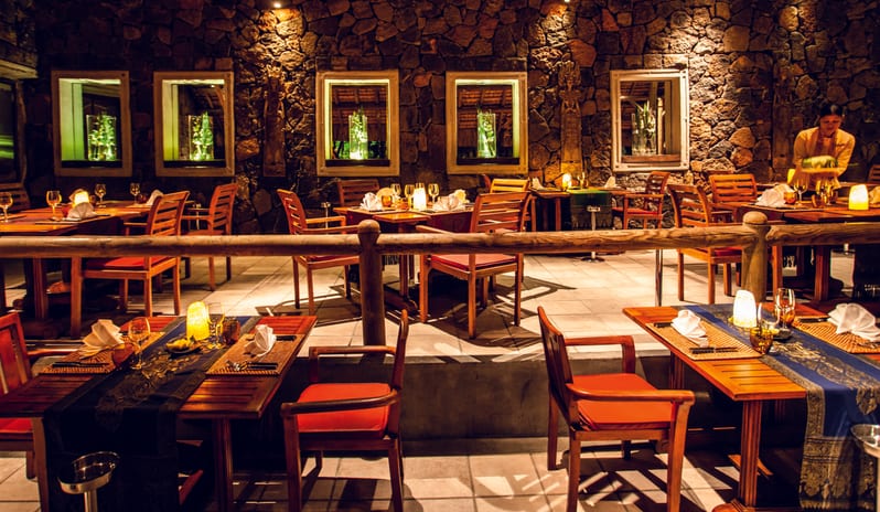Teak Elephant Fine dining restaurant dining room with elegant table settings, glowing candles, blue table runners, and a server attending to guests in the background. at Shandrani for 2 by Kenwood Travel