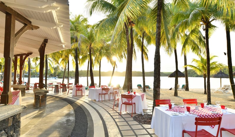 Ponte Vecchio Seaside restaurant tables on a paved patio overlooking a tropical beach with palm trees at Shandrani for 2 by Kenwood Travel