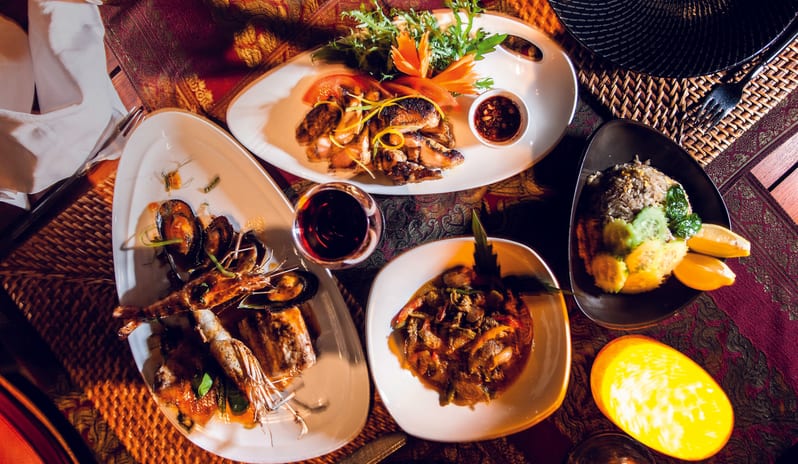 Teak Elephant various traditional dishes on elegant white and black plates, a glass of red wine, textured purple tablecloths, and warm ambient light at Shandrani for 2 by Kenwood Travel