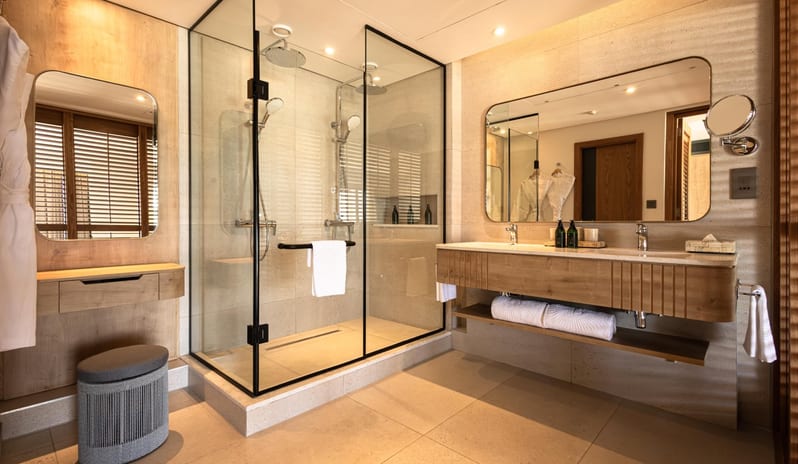 Bay View Room A modern walk-in glass shower enclosure featuring dual rain showerheads and handheld sprayers, paired next to a light wood vanity and makeup station at Shandrani for 2 by Kenwood Travel