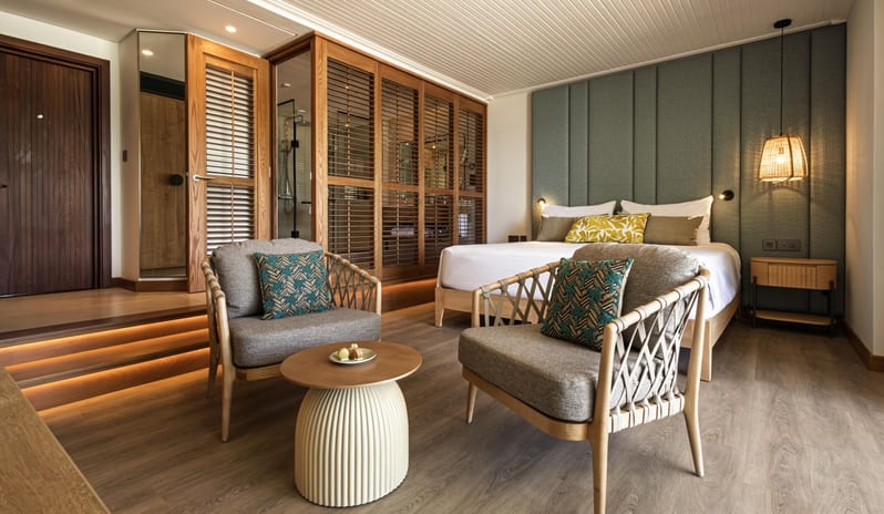Bay View Room king bed with a green upholstered headboard, wooden louvered partitions, and a stylish sitting area with woven armchairs at Shandrani for 2 by Kenwood Travel