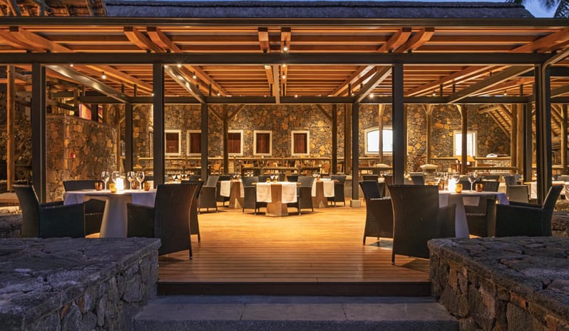 Serendipity (Adult-only restaurant) An upscale, open-air resort restaurant at night featuring set dining tables on a wooden deck, stone accent walls, and warm overhead lighting at Shandrani for 2 by Kenwood Travel