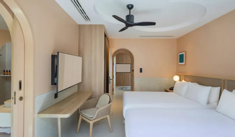 Two Bedroom Pool Villa two twin beds with white linens, a wall-mounted TV over a wooden desk, a wicker chair, and an arched doorway leading to an ensuite bathroom at Centara Karon Villas Phuket by Kenwood Travel
