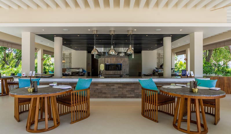 Med Modern open-air resort restaurant featuring a stone pizza oven, marble-top tables with teal cushions, and tropical garden views at Kuda Villingili Resort Maldives by Kenwood Travel