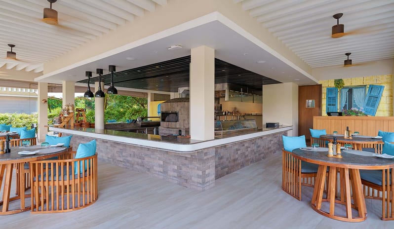 Med Modern open-air resort restaurant featuring a stone pizza oven, marble-top tables with teal cushions, and tropical garden views at Kuda Villingili Resort Maldives by Kenwood Travel