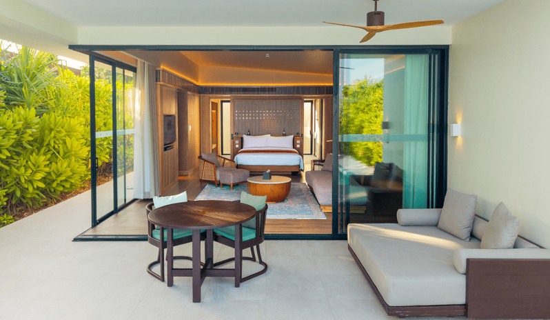 Sunrise Beach Villa with Private Pool View from a private terrace of a luxury beach villa, showing an outdoor daybed and dining set leading into a modern wood-paneled bedroom at Kuda Villingili Resort Maldives by Kenwood Travel