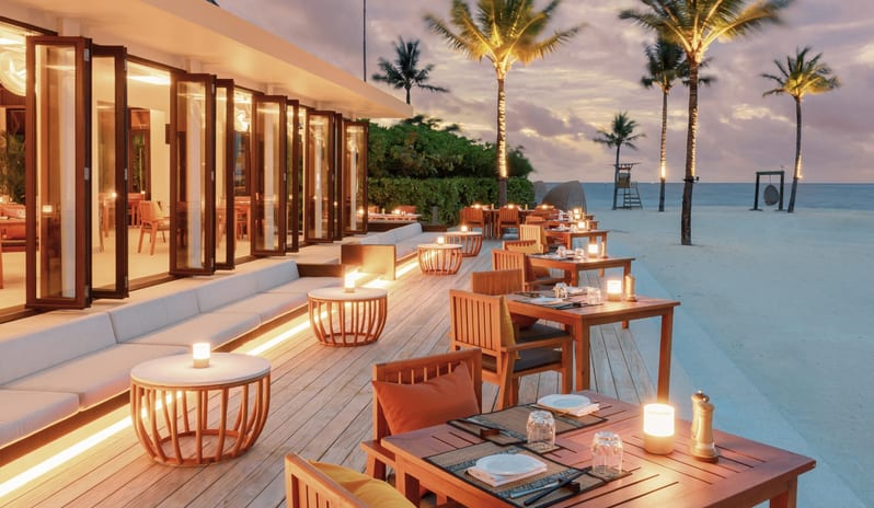 Mar-Umi A luxury beachfront restaurant patio with wooden tables, soft warm lighting, and a view of the ocean at sunset at Kuda Villingili Resort Maldives by Kenwood Travel