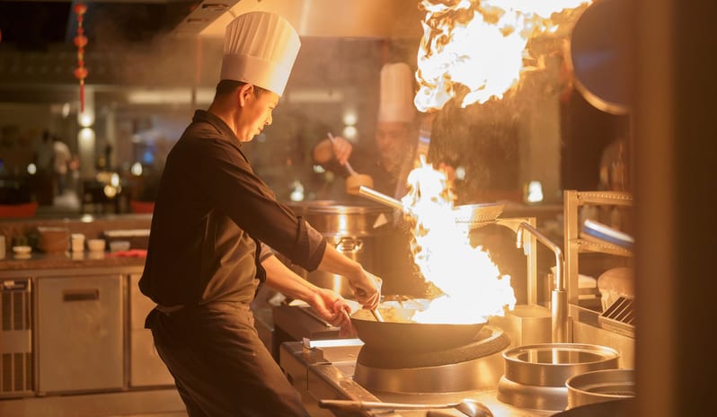 East A professional chef in a black uniform and tall white toque cooking with a large flame in a wok at a luxury resort's open kitchen at Kuda Villingili Resort Maldives by Kenwood Travel