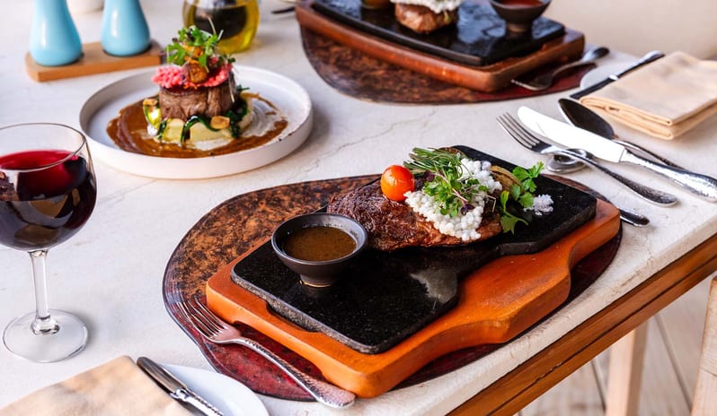 Fire Gourmet steak served on a black stone platter with micro-greens and jus, paired with a glass of red wine on a white stone table at Kuda Villingili Resort Maldives by Kenwood Travel