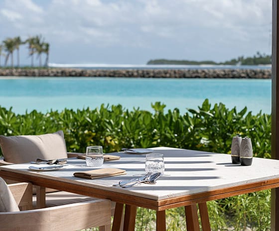 The Restaurant Close-up of a set outdoor dining table with stone top and wooden chairs on a deck, overlooking a bright blue lagoon and tropical island greenery under a sunny sky at Kuda Villingili Resort Maldives by Kenwood Travel