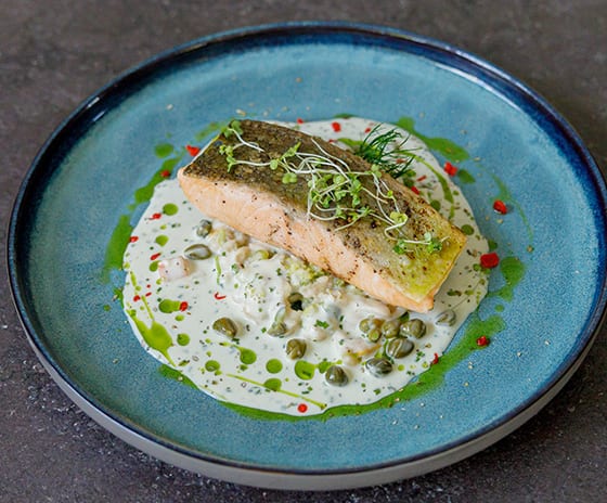 Med Pan-seared salmon fillet served over a creamy white sauce with capers, herb oil drizzles, and microgreen garnish on a blue ceramic plate at Kuda Villingili Resort Maldives by Kenwood Travel