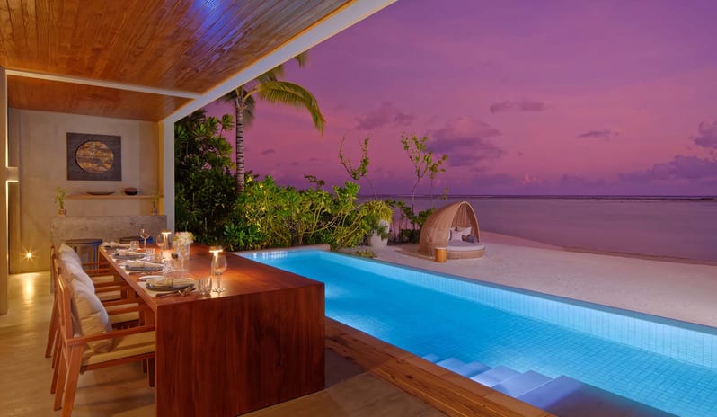 Three bedroom Beach Retreat with Private Pool Al fresco evening dining area with a long wooden table next to a lighted private lap pool at a luxury beach villa during a purple sunset at Kuda Villingili Resort Maldives by Kenwood Travel