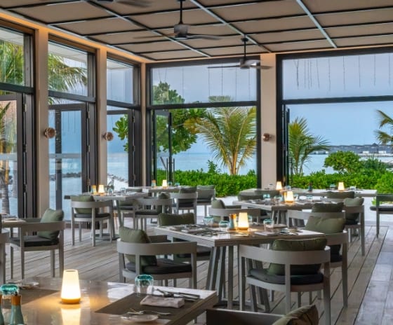 The Restaurant modern resort restaurant with floor-to-ceiling glass doors open to a tropical beach, featuring grey minimalist chairs, wooden flooring, and warm table lamps at Kuda Villingili Resort Maldives by Kenwood Travel