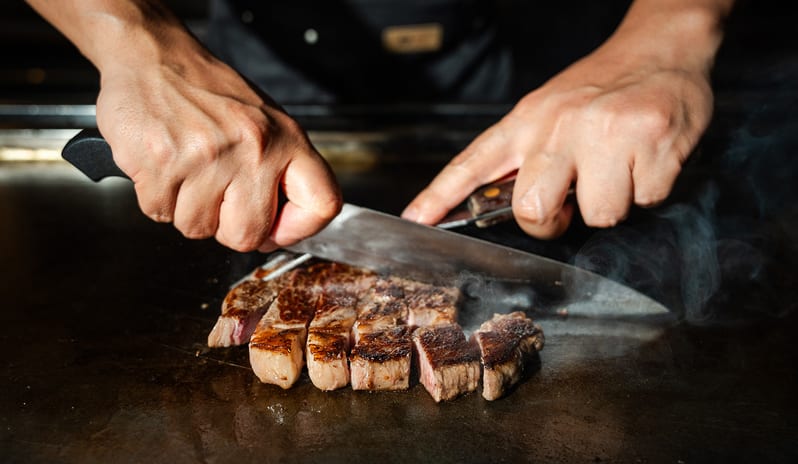 Teppanyaki by Mar-Umi Close-up of a chef’s hands slicing seared wagyu beef steak on a steaming teppanyaki grill at Kuda Villingili Resort Maldives by Kenwood Travel