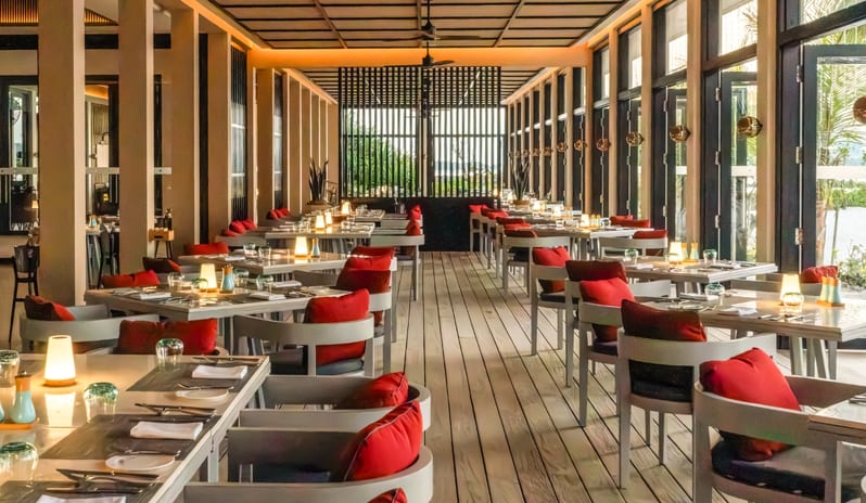 Fire Interior of a modern fine-dining restaurant featuring white wooden tables, red cushions, and large floor-to-ceiling windows overlooking a tropical coast at Kuda Villingili Resort Maldives by Kenwood Travel