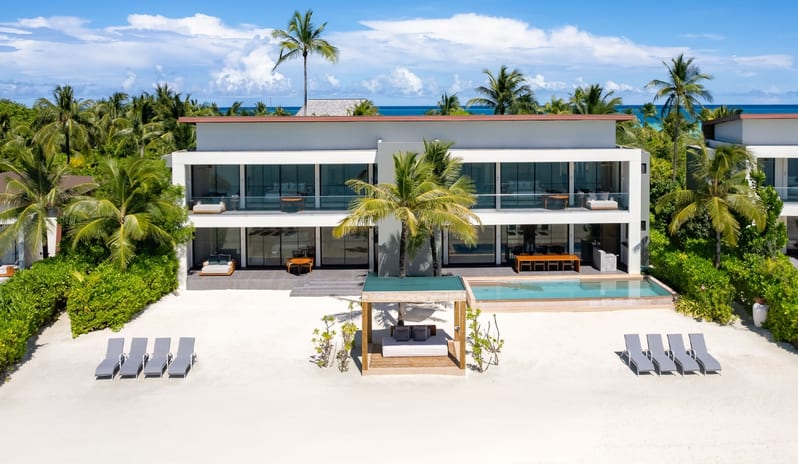 Three bedroom Beach Retreat with Private Pool Exterior view of a modern two-story three-bedroom beach retreat with a private pool, beachfront lounge chairs, and tropical palm trees under a blue sky at Kuda Villingili Resort Maldives by Kenwood Travel