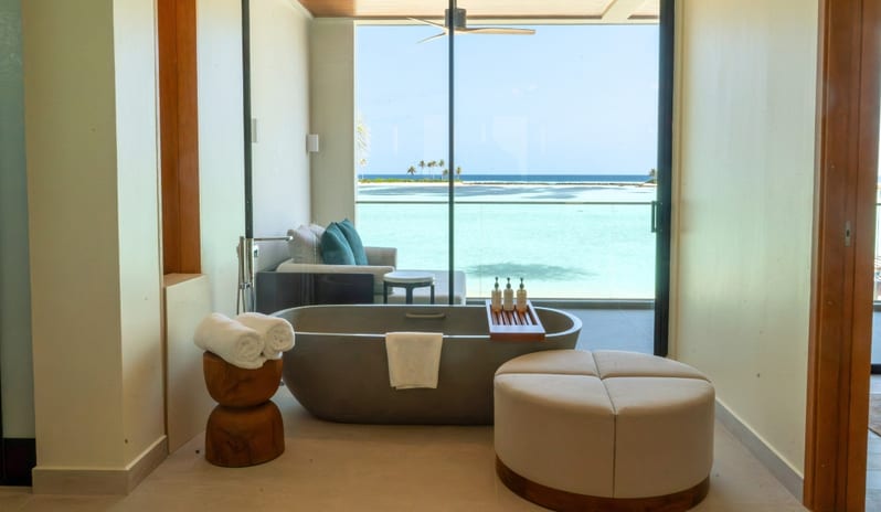 Three bedroom Beach Residence with Private Pool stone freestanding bathtub, a circular ottoman, and floor-to-ceiling windows overlooking a turquoise ocean lagoon at Kuda Villingili Resort Maldives by Kenwood Travel