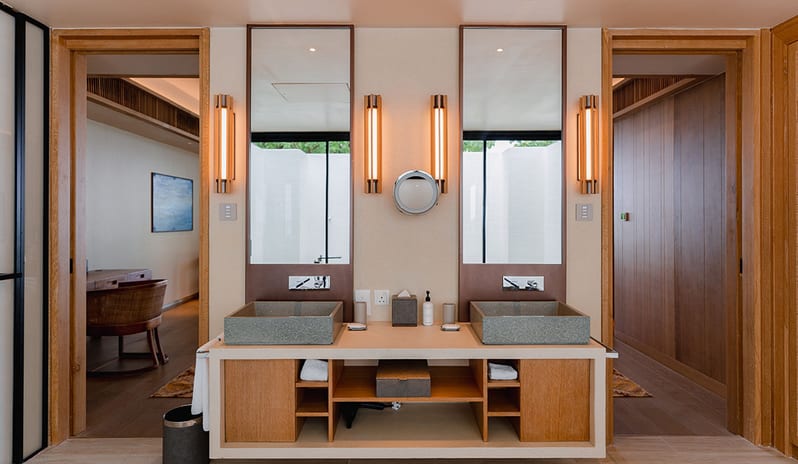 Sunset Water Villa with Private Pool two grey stone vessel sinks, tall vertical sconce lighting, and large rectangular mirrors, flanked by wooden doorways leading to a walk-in closet and bedroom at Kuda Villingili Resort Maldives by Kenwood Travel